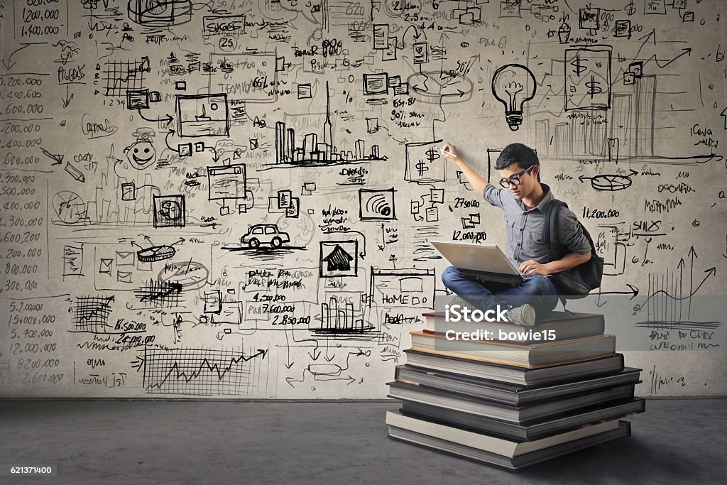 Young Asian boy with nerdy black glasses sitting on a book hill, drawing his ideas creatively on the wall behind him while looking at his computer.